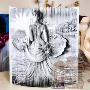 Op de afbeelding: Een zwart-witte boekkunst-sculptuur van een vrouw die op een strand loopt met haar jurk die achter haar waait. De sculptuur is gemaakt van pagina's van een boek die zijn gevouwen en geknipt om de afbeelding te creëren.