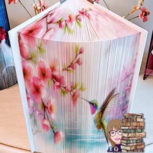 Może przedstawiać: Rzeźba z książki złożonej w kształt kolibra i różowych kwiatów na białym tle. Rzeźba wykonana jest ze stron książki i jest unikalnym i pięknym dziełem sztuki.