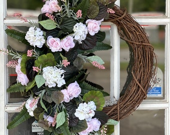 May include: A grapevine wreath adorned with faux pink roses, white hydrangeas, and green foliage. The wreath is decorated with sprigs of pink flowers and dark green leaves, creating a natural and elegant look. The wreath is hanging on a white door.