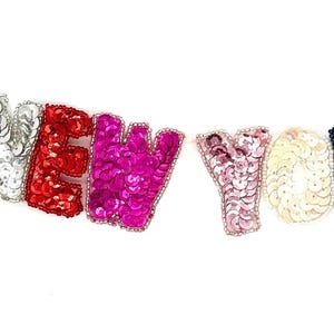May include: A banner spelling "NEW YORK" in individual sequined letters. The letters are in various colors, including silver, red, pink, light pink, dark teal, and green, against a white background. The sequins are outlined with a beaded trim.