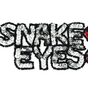 May include: A decorative applique with the words "SNAKE EYES" spelled out in silver sequins, outlined with black beads. Two red sequined diamond shapes are on the right side of the text. The applique is on a white background.