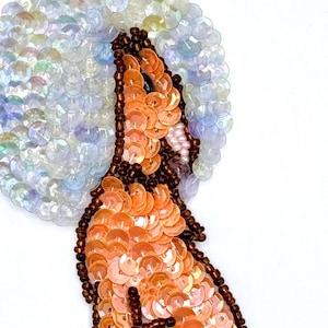 May include: A brown sequined wolf howling at a white iridescent sequined moon. The wolf is sitting on a bed of brown and beige sequins.