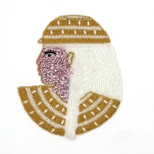 May include: An embroidered applique featuring a profile of a person with a pink sequined face, a gold beaded headdress, and white beaded hair. The design includes gold beaded accents and a fan-shaped collar.
