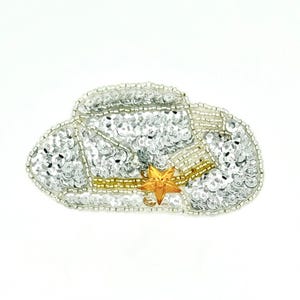 May include: A silver sequined cowboy hat patch featuring a gold star accent on the hatband. The patch is embellished with intricate silver and gold beadwork along the edges and crown, creating a textured, reflective surface in a western shape.