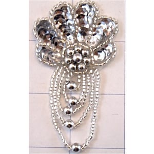 May include: A silver-toned, beaded applique with a floral design. The applique features a central flower shape with a cluster of small beads, surrounded by larger, reflective sequins. Dangling beaded strands with silver beads complete the design.
