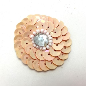 May include: A circular floral embellishment made of peach-coloured sequins. The centre features a large, iridescent gem surrounded by small white beads. This decorative item is suitable for crafts and embellishments. The sequins are arranged in a petal-like pattern.