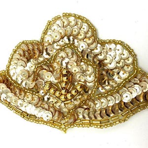 May include: A gold sequin applique in a floral design. The applique is heavily embellished with gold sequins and small gold beads, creating a textured and ornate appearance. The design is symmetrical and detailed.