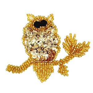 May include: A decorative owl patch crafted with gold-toned seed beads and metallic sequins. The owl features two prominent black circular beads for eyes and is perched on a branch made of matching gold beads, creating a textured, shimmering effect.