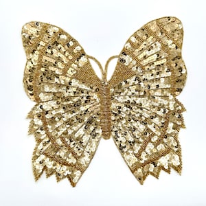 May include: A gold sequined butterfly applique. The butterfly is made of small, round sequins that are sewn together to create a shimmering effect. The butterfly has a detailed design with a central body and two wings.