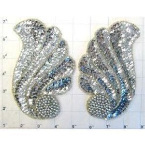 May include: Two silver sequined applique patches in a stylized feather or wing design. Each patch features a curved, layered pattern of sequins, with a rounded base. The patches are approximately 6 inches tall.