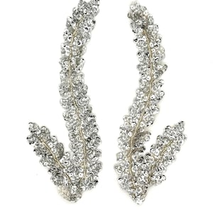 May include: A pair of silver sequin appliques shaped like curved leafy branches. Each piece features dense, reflective silver sequins stitched onto a base, creating a textured, shimmering appearance with a distinct stem and branching foliage.