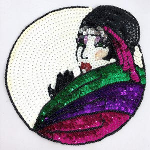 May include: A round, sequined patch featuring a stylized face. The design incorporates white, black, green, purple, and pink sequins. The face is outlined with black beads, and the background is white. The patch is likely for decorative purposes.