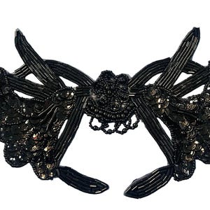 May include: A black decorative applique featuring intricate beadwork and sequins. The design includes elongated, tube-shaped beads arranged in radiating patterns, accented by clusters of round, shimmering sequins and a central beaded floral motif.