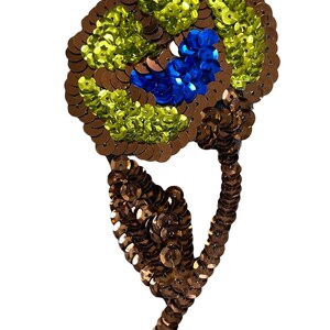 May include: A sequined patch shaped like a flower with a stem and leaf. The flower head features concentric circles of brown, bright lime green, and a central cluster of royal blue sequins. The stem and leaf are covered in brown sequins.