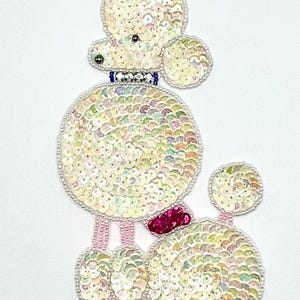 May include: A white sequined poodle with a pink bow and a blue collar. The poodle is standing on its hind legs with its front paws raised. The poodle is surrounded by white sequins.