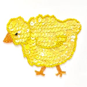 May include: A decorative patch shaped like a small chick, crafted with bright yellow sequins and beads. The chick features an orange beaded beak and two orange beaded feet, with a single small black bead serving as the eye on its head.