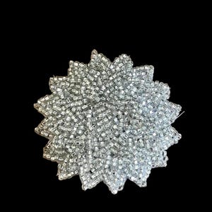 May include: A circular beaded applique featuring a dense arrangement of small, reflective silver-toned glass beads. The piece is shaped like a stylized flower or star with multiple pointed petals radiating from the center, creating a textured surface.