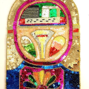 Peut inclure: Un patch en sequins coloré représentant un juke-box vintage avec un cadre doré, une base rose et bleue et un motif coloré au centre.