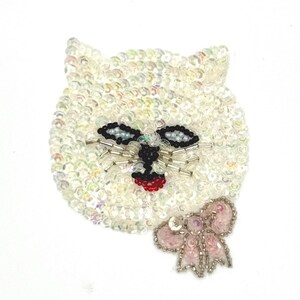 May include: A cat-shaped patch covered in iridescent white sequins. The face features black beaded eyes, silver tube bead whiskers, and a red beaded nose. A small, light pink sequined bow with silver bead accents is attached to the lower right side.
