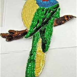 May include: A colorful sequined parrot perched on a brown branch. The parrot features red, green, yellow, and blue sequins, with a white eye and a green head. The branch is brown with brown sequins. The background is white.