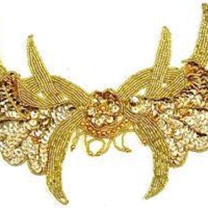 May include: A gold-colored, beaded applique with a symmetrical design. The applique features a central floral element, two wing-like sections with sequin details, and multiple upward-curving, antler-like extensions. The design is set against a white background.