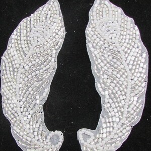 May include: Two white beaded feather-shaped appliques with silver accents. The appliques are on a white mesh fabric.