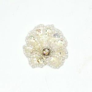 May include: A circular floral applique crafted from clear and iridescent white beads. The design features a central faceted bead surrounded by layers of smaller seed beads and larger, reflective crystal-like accents arranged in a petal pattern.