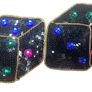 Peut inclure: Deux dés noirs avec des bordures dorées et des paillettes colorées. Chaque dé a un motif différent de paillettes roses, bleues et vertes.