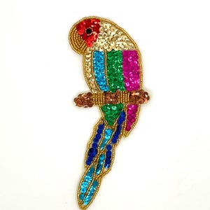 May include: A vibrant sequined parrot applique. The parrot has a red head, gold body, and a tail with blue, green, and gold sequins. It is perched on a brown sequined branch. Ideal for embellishing clothing or craft projects.