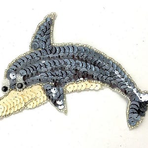 May include: A dolphin-shaped patch crafted with dark gray and cream-colored sequins. The body is covered in dark gray sequins, while the underside and snout feature cream-colored sequins. The edges are outlined with small, clear glass beads.