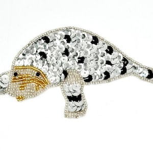 May include: A decorative patch shaped like a manatee, crafted with silver and black sequins. The head features gold-toned beadwork for the snout and whiskers, with small black beads for the eyes. The entire outline is bordered by tiny silver beads.