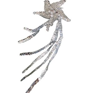 May include: A silver star-shaped appliqué covered in shimmering sequins and beads. The star features five points and is adorned with five long, trailing strands of silver sequins that extend downward, creating a cascading, comet-like effect.