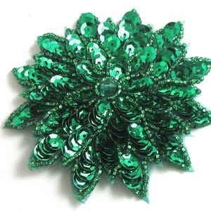 Peut inclure: Une embellissement de fleur en paillettes vertes avec une finition brillante. La fleur est faite de paillettes et de perles superposées, créant un aspect texturé et dimensionnel.