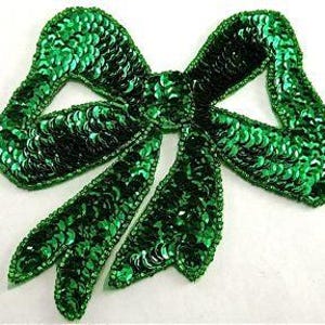 May include: A green sequined bow applique. The bow is made of small, round sequins that are sewn together to create a three-dimensional shape. The bow is perfect for adding a touch of sparkle to any project.