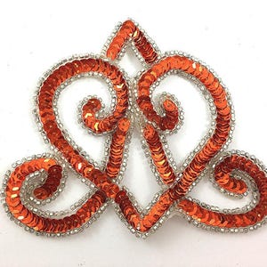 May include: An orange sequin applique in a heart-shaped design with a silver beaded border. The decorative embellishment features a symmetrical pattern with swirling details, perfect for adding flair to clothing or accessories.