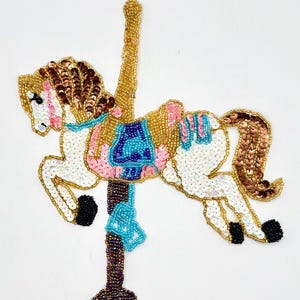 May include: A white and brown beaded carousel horse with a blue, pink, and gold saddle. The horse is attached to a brown beaded pole.