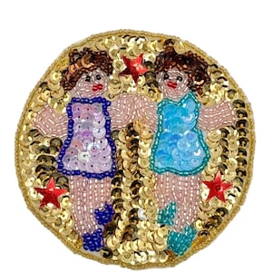 May include: A round, beaded brooch featuring two figures in dresses, surrounded by gold sequins. The figures are in blue and purple dresses, with red star accents. The brooch is approximately 3 inches in diameter.