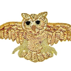 May include: A decorative owl patch featuring gold sequins and beads. The owl is depicted with wings fully spread, a textured body, and two prominent black circular eyes. The entire surface is covered in metallic gold embellishments.