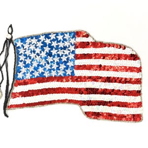 May include: A sequined American flag design with red and white stripes and a blue field of white stars. The flag is embellished with silver beads around the edges and has a black beaded string with decorative accents.