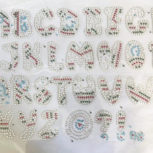 May include: Alphabet letters A through Z, each crafted with silver-colored rhinestones. The letters are embellished with red, green, and blue accents, creating a sparkling, decorative design. The letters are arranged on a white surface.