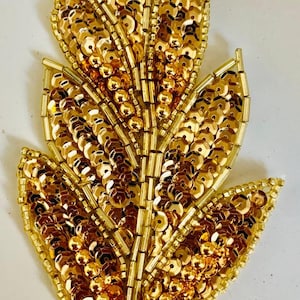 May include: A gold sequin leaf applique. The leaf is made of gold sequins and beads, arranged in a leaf shape. The applique is likely for embellishing clothing or accessories.