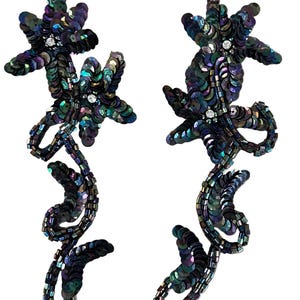 May include: A pair of long, dangling earrings featuring iridescent dark purple and blue sequins arranged in a floral and vine-like pattern. Each earring includes small clear crystals at the center of the flower shapes and a curved, trailing stem design.