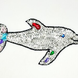 May include: A dolphin-shaped patch covered in silver sequins with a black beaded outline. The design features colorful teardrop-shaped rhinestones in red, green, blue, purple, and light pink accents on the fins and tail, with a small blue eye.