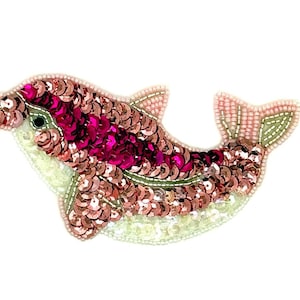 May include: A dolphin-shaped patch crafted with metallic pink and magenta sequins. The body features a gradient of pink sequins, while the underbelly is detailed with white beads and sequins. A single black bead serves as the eye of the dolphin.