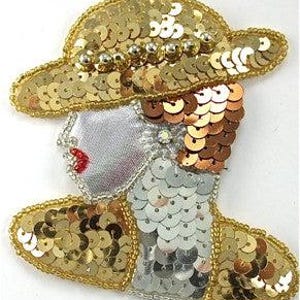 May include: A gold sequined applique in the shape of a woman wearing a hat. The hat and the woman's shoulders are adorned with gold sequins. The woman's face is silver with red lips.