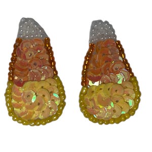 Peut inclure: Une paire de boucles d'oreilles pendantes en forme de maïs sucré. Les boucles d'oreilles sont faites de sequins orange, jaune et blanc irisés.