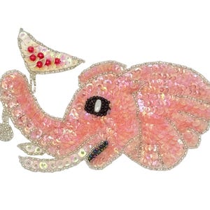 May include: A pink sequined elephant-shaped applique with a silver beaded outline. The elephant has a black eye and a small white tusk. It holds a white flag with red accents. The applique is likely for clothing or accessories.