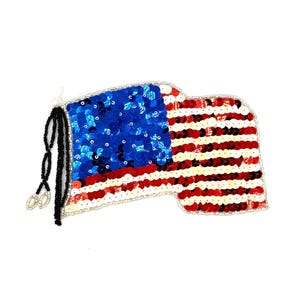 May include: A sequined American flag design, featuring a blue field with white stars and red and white stripes. The flag is embellished with sequins and has a black beaded tassel. The design is on a white background.