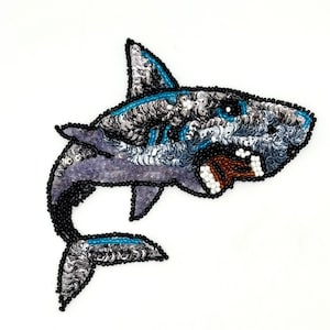 May include: A shark-shaped patch crafted with silver, blue, and black sequins and beads. The design features a shark with an open mouth showing white teeth and a brown interior, outlined in black beads against a plain white background.