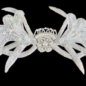 May include: A white, beaded headpiece with a floral and crown design. The headpiece features multiple curved, petal-like elements and is embellished with sequins and beads. The central design includes a small crown and a flower.